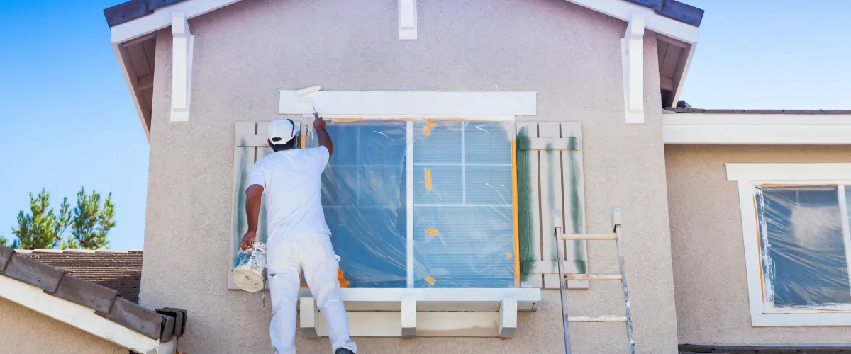 A house painter in white clothing standing on a ladder, using a roller to paint the exterior window trim of a beige stucco house.