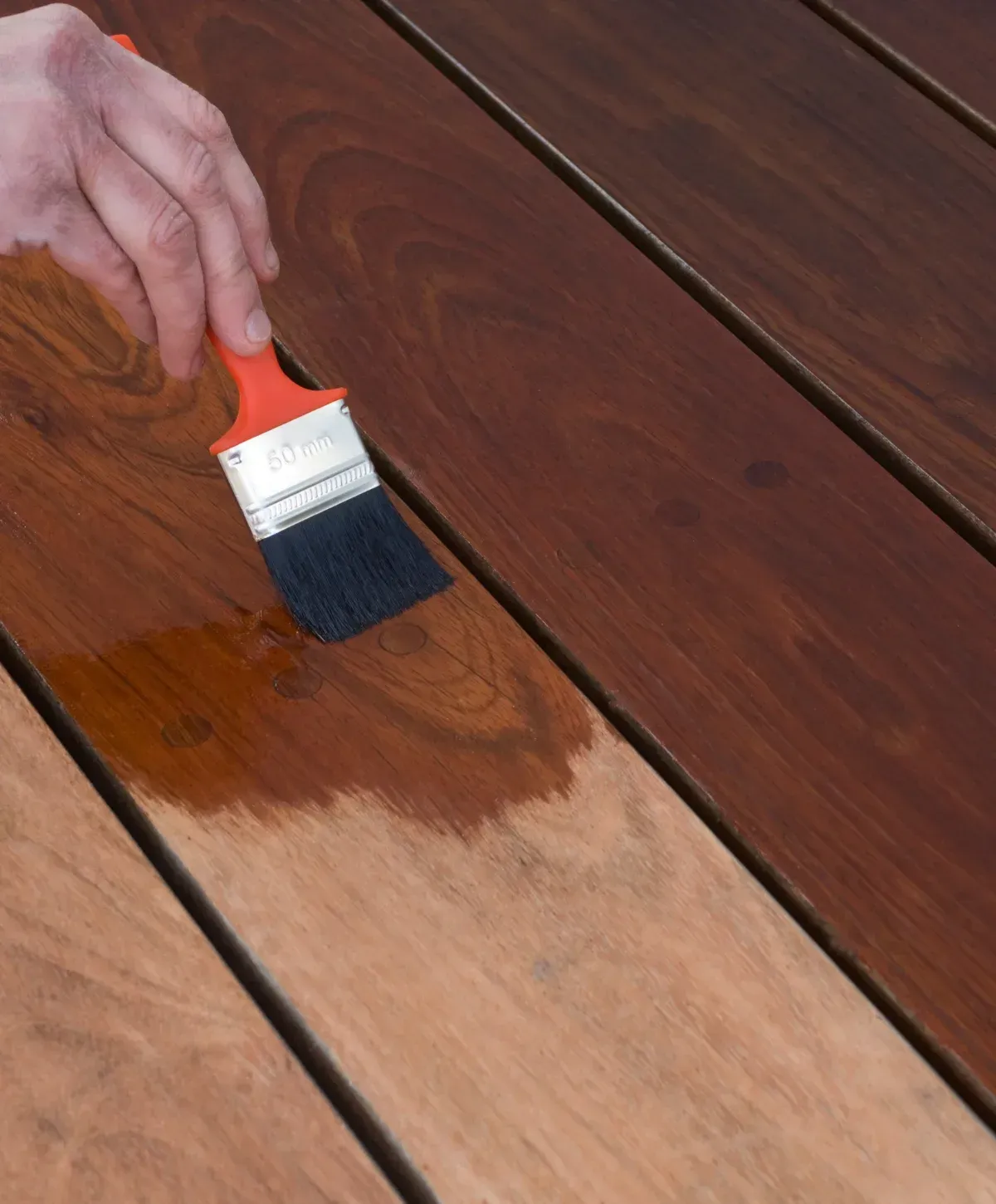A person in green gloves applies brown wood stain to outdoor deck planks with a large brush on a sunny day.