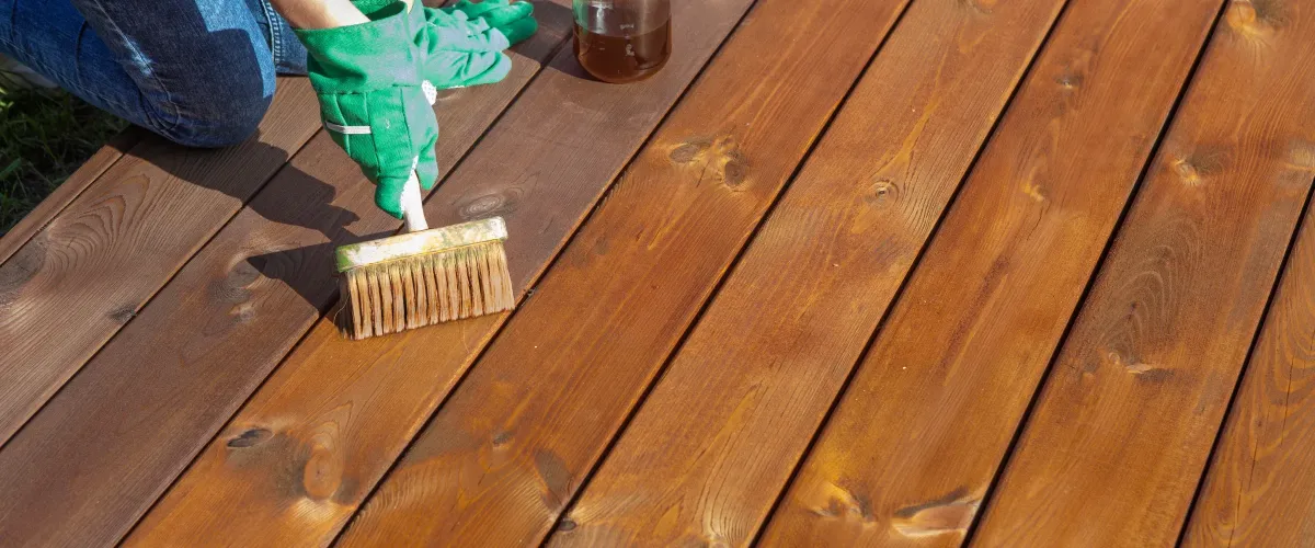 A hand holds an orange-handled brush, applying dark wood stain to a light wooden deck board, showing a half-finished surface.
