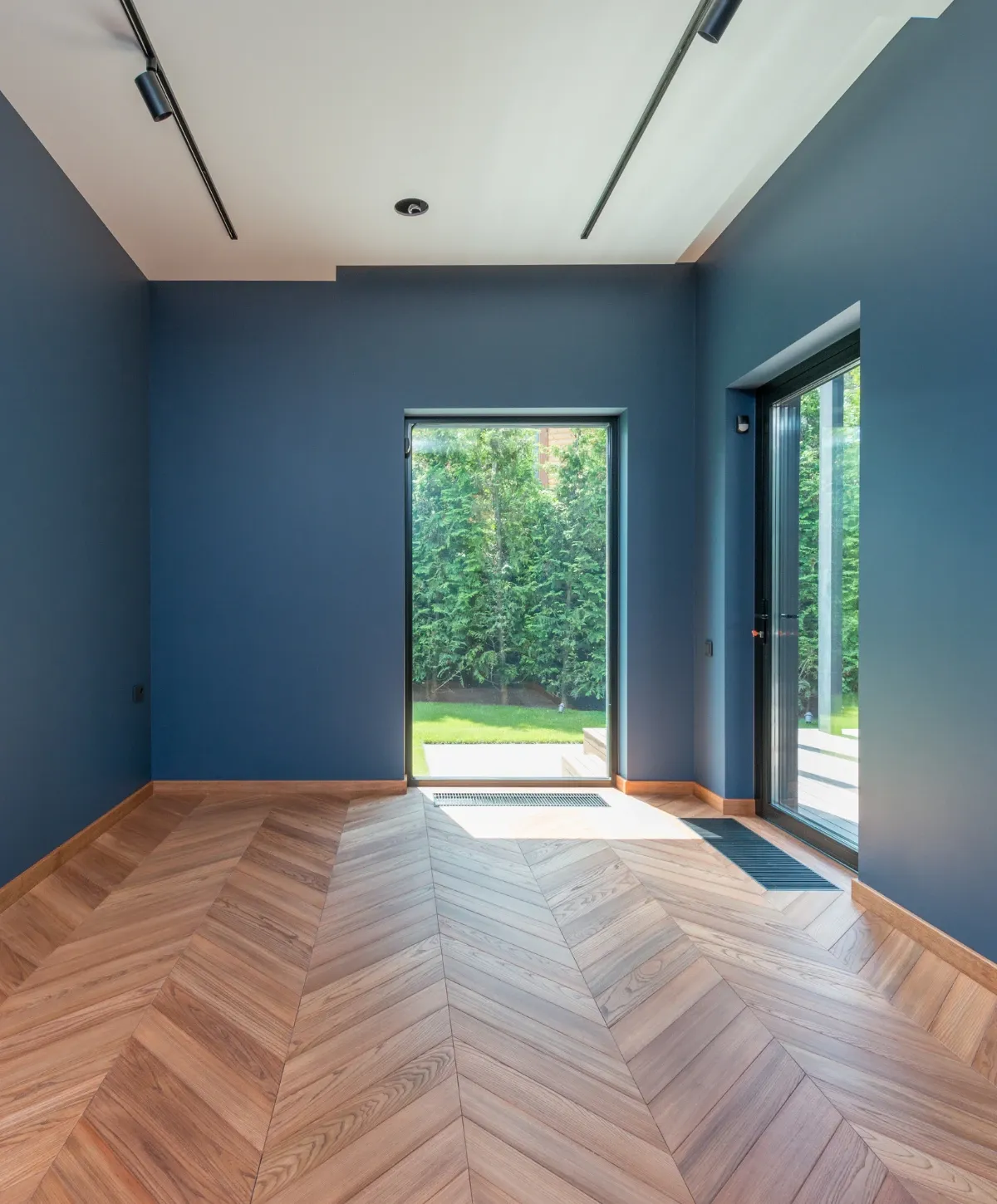 An empty modern room featuring dark blue walls, light herringbone wood flooring, and two tall glass doors opening to a green garden.