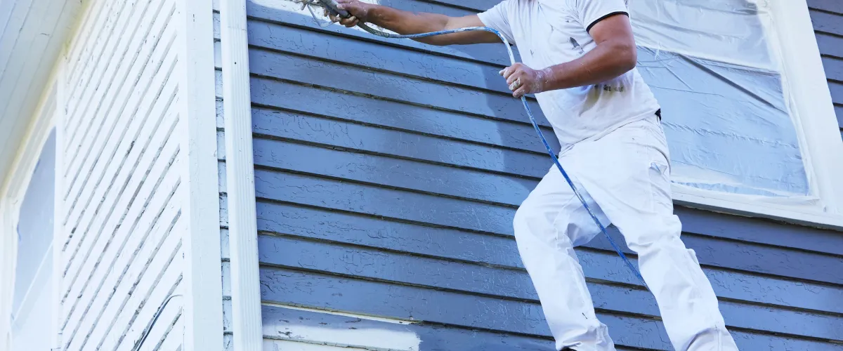 A person in white overalls uses a spray gun to paint blue siding on a house, with a protected window visible.