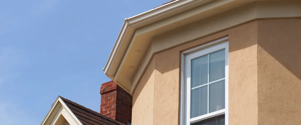 A close-up view of a house exterior with beige stucco, white window, gutters, and a brick chimney against a blue sky.