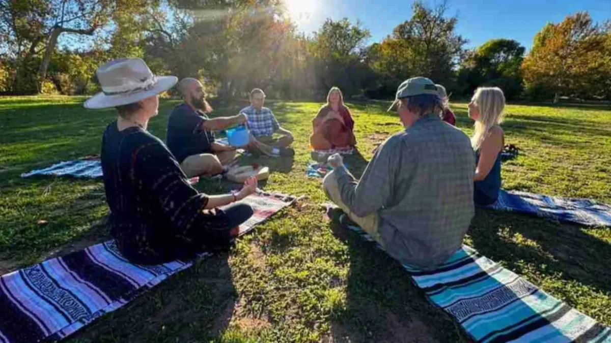 Group circle sitting on blankets, resting on green grass, surrounded by trees. 7 people engaged in breathwork.