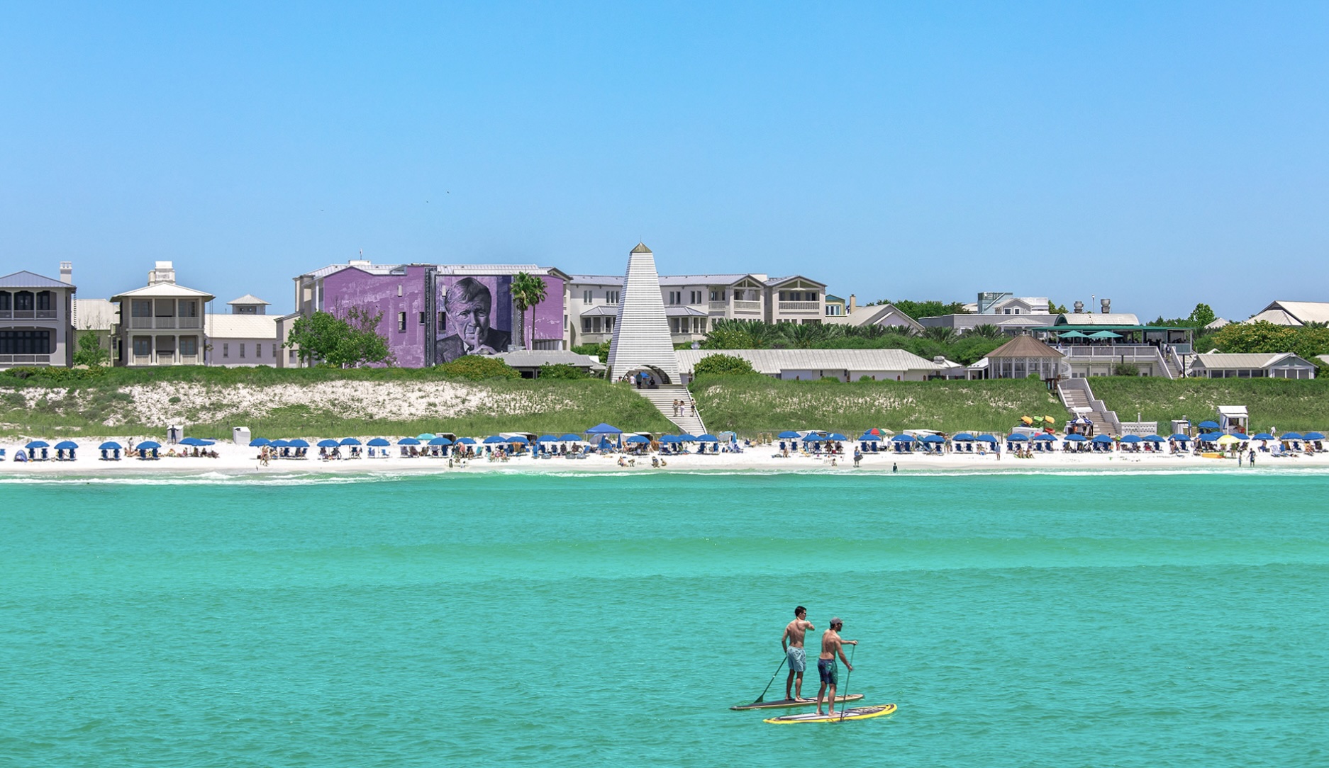 Aerial view of Seaside, Florida with turquoise Gulf waters, white sand beach, colorful beachfront buildings, and people paddleboarding offshore.