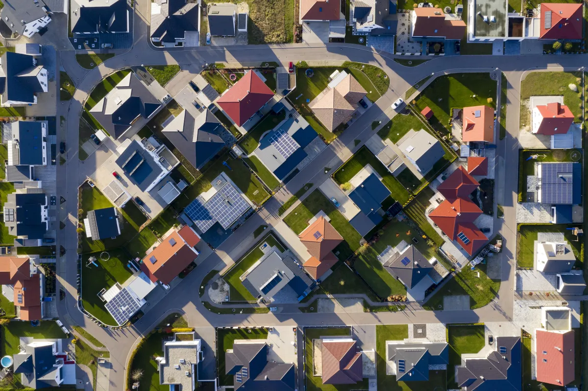 An overhead aerial photograph of a suburban neighbourhood, showing rows of detached houses with red and dark grey roofs, curving streets, driveways, green lawns and solar panels on several roofs.
