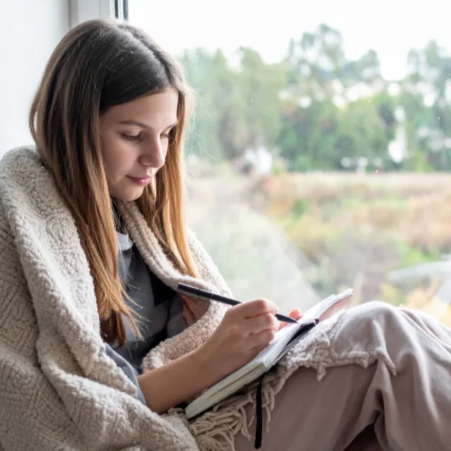 Teenager sitting by a window writing in a journal, using reflection and journaling as a strategy to understand and regulate emotions.