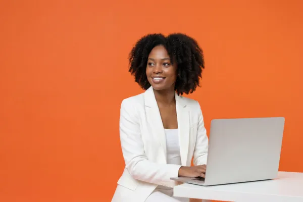Woman looking to the right with laptop open and hands on the laptop, background is orange and woman wearing white.