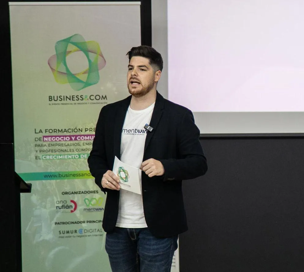 Jorge T., a young Latino man with glasses, short dark hair, wearing a teal shirt, holding a microphone, mid-speech, soft orange background. His expression is animated and passionate, engaging the audience with every word.