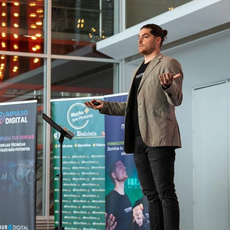 Jorge T., a young Latino man with glasses, short dark hair, wearing a teal shirt, holding a microphone, mid-speech, soft orange background. His expression is animated and passionate, engaging the audience with every word.