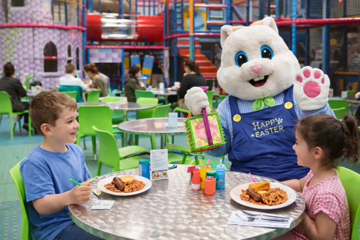 A cheerful Easter Bunny in a pastel costume waves beside a breakfast table set with pancakes, fruit, and juice, surrounded by smiling children of diverse backgrounds in spring attire, inside a sunlit, festively decorated amusement centre.