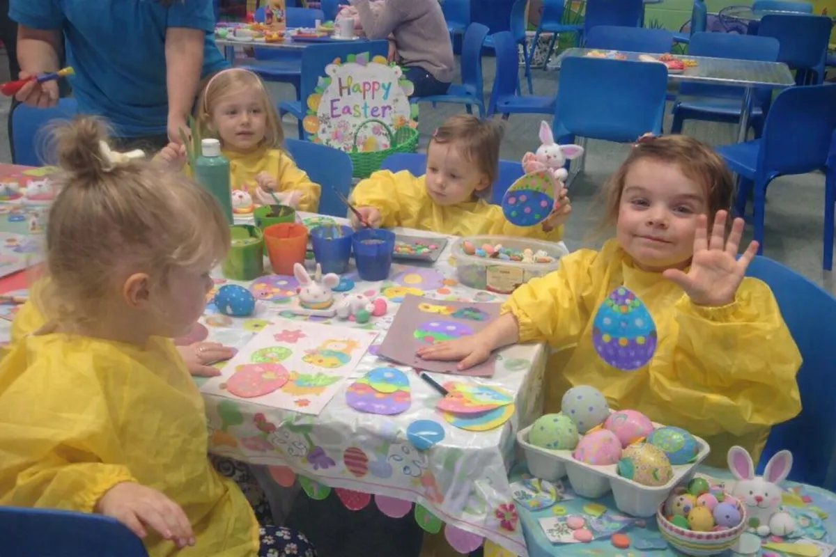 Children of various ages and backgrounds gather around a large table, joyfully painting colorful Easter eggs with brushes and stickers, while a parent assists, in a bright, lively room decorated with streamers and bunny motifs.