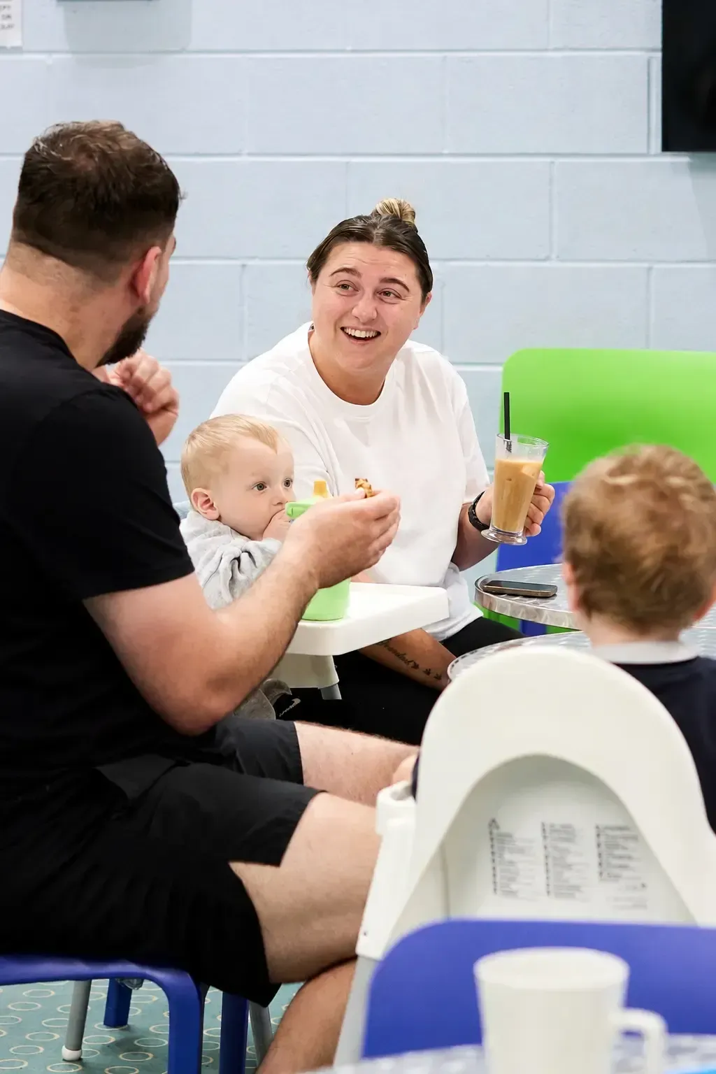 A candid, brightly lit café scene capturing a cheerful family moment. A smiling mother with a top bun and a small arm tattoo enjoys an iced coffee while engaging with a bearded father in black, who holds a snack. Two young children sit nearby in white high chairs, one with a green sippy cup. Colorful seating and a pale blue cinderblock wall create a lively, modern children’s area atmosphere.
