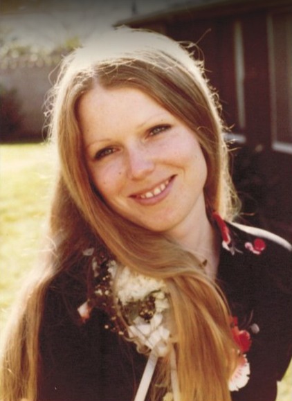 Dot Harvey speaking at a community event, standing at a podium with attentive listeners in the background. She is animated and expressive, wearing a patterned dress, and the setting is a warmly lit hall decorated with banners and flowers.