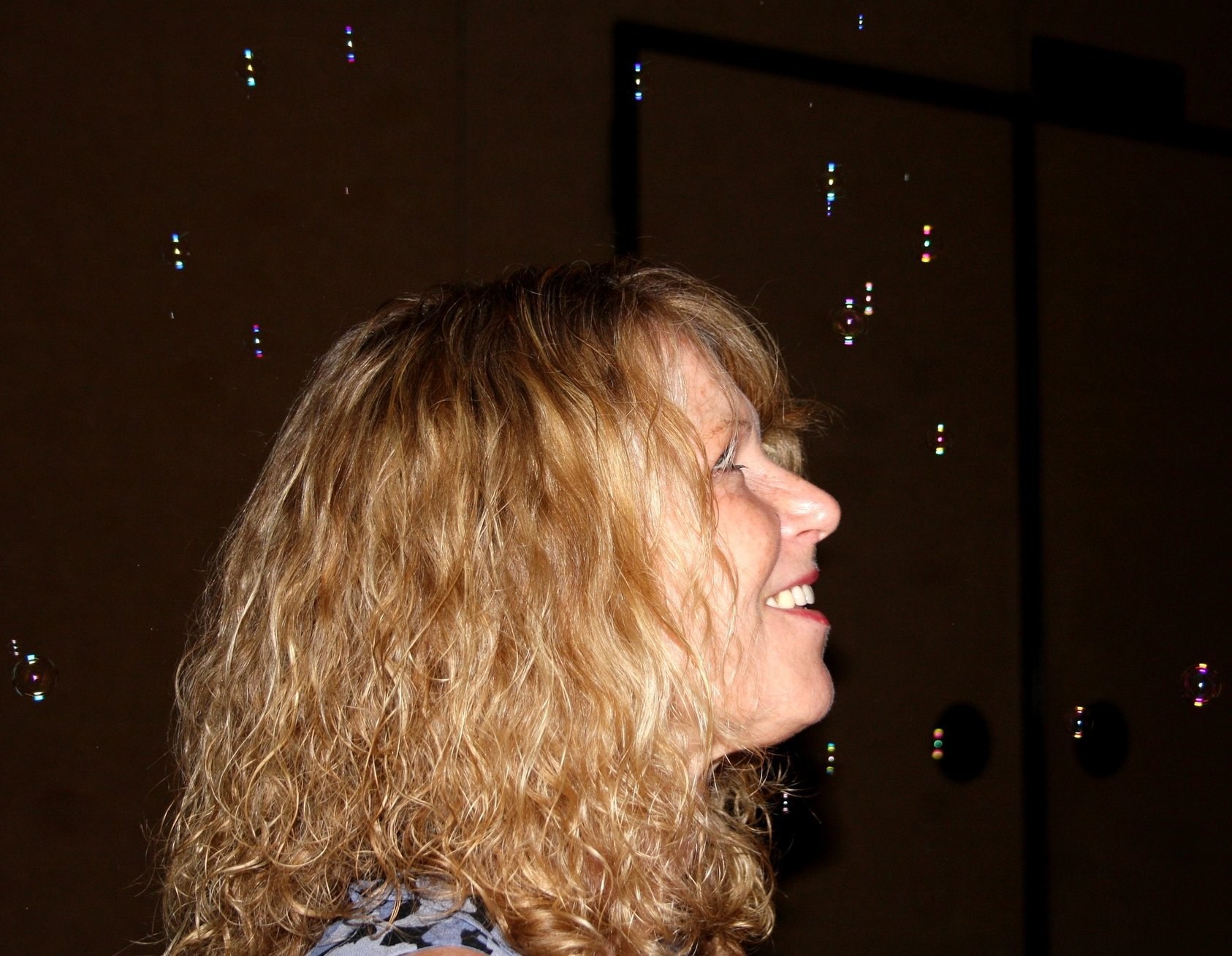 Dot Harvey tending to her vibrant garden, kneeling among colorful flowers and lush plants. She wears a straw hat and gardening gloves, her face lit with a gentle smile. The scene is bright and lively, showcasing her passion for nature and nurturing life.