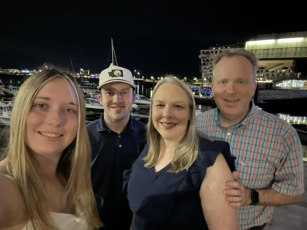 A candid photo of the Nelson family gathered on a sunlit porch, laughing together. The group includes two adults and three children of diverse ages, all dressed in casual, colorful clothing. The background features lush greenery and a wooden swing, evoking warmth and togetherness.