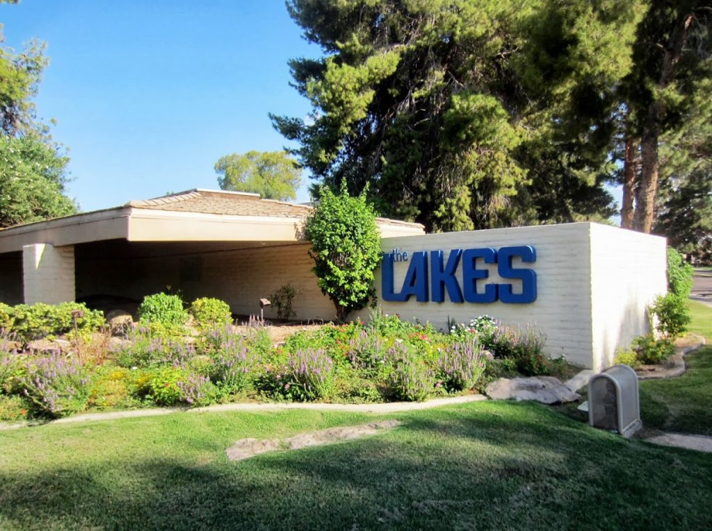 An exterior view of Tempe Lakes Tennis & Beach Club, showing the entrance with clear signage, palm trees, and a welcoming pathway. The sky is bright blue, and the setting feels inviting and accessible for all visitors.