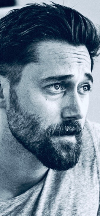 A young Latino man with a friendly smile, wearing a Modern Breath instructor badge, sits cross-legged on a yoga mat in a bright studio. The background features soft, neutral tones and subtle yoga props, conveying expertise and warmth.