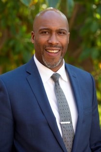 A confident coach in a navy blazer, standing in front of a whiteboard filled with strategy diagrams, smiling warmly. The setting is a bright, professional workspace, with subtle gold and blue accents, conveying expertise and approachability.