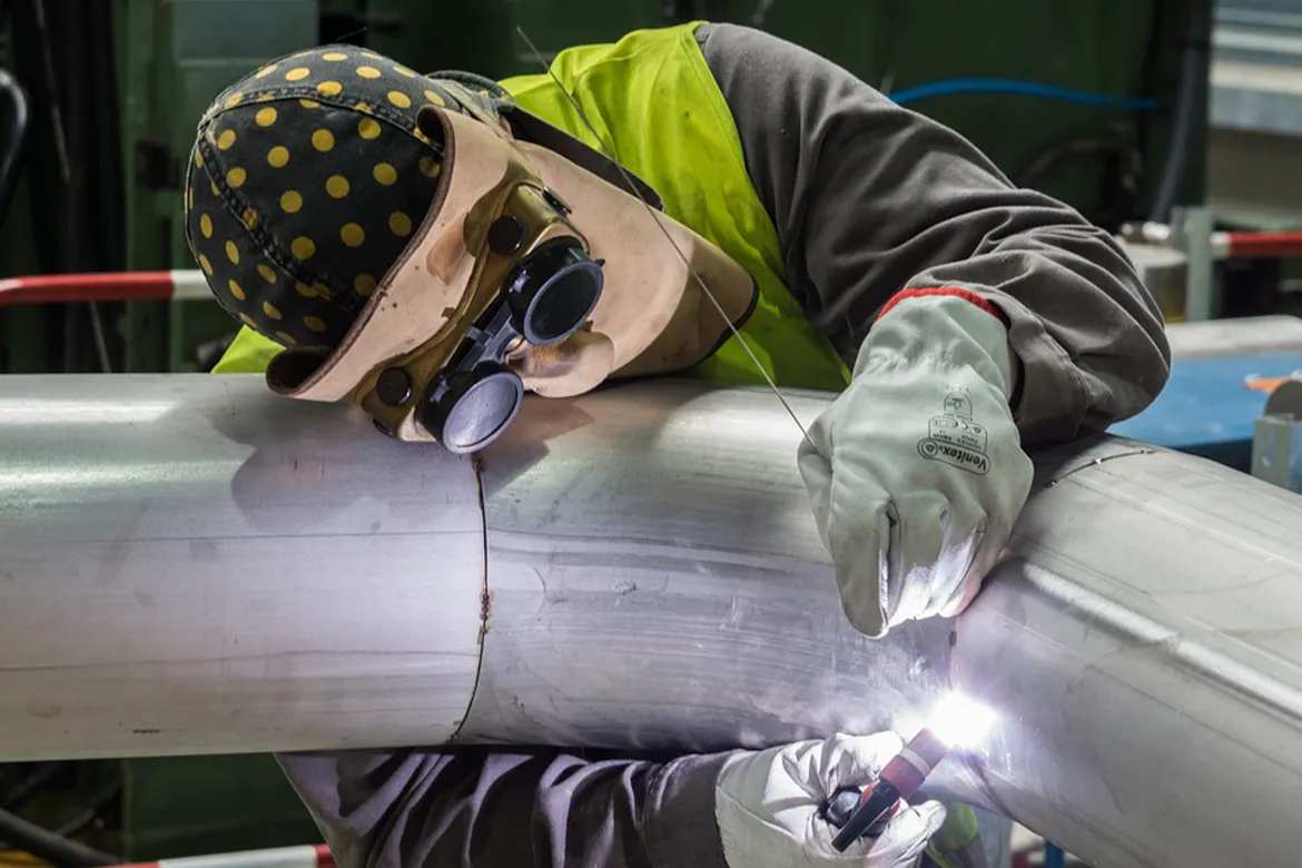 Industrial worker welding large metal pipes while wearing protective gloves, mask, and safety gear.