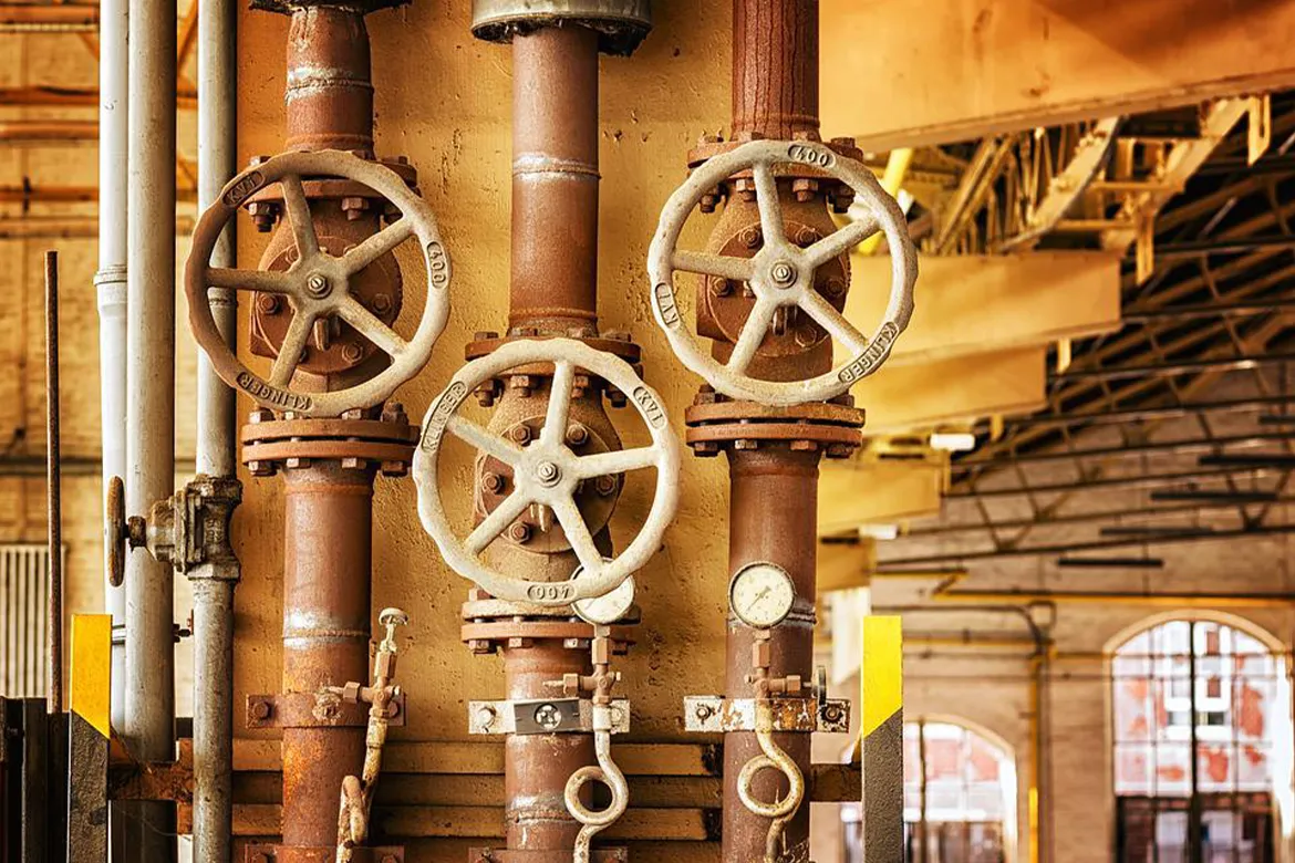 Old industrial plumbing system with large rusted pipes, valve wheels, and a pressure gauge inside a factory building.