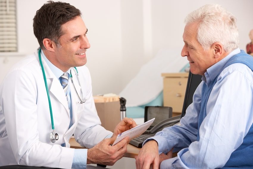A male doctor in a white coat smiling and consulting with an elderly male patient in a clinic setting