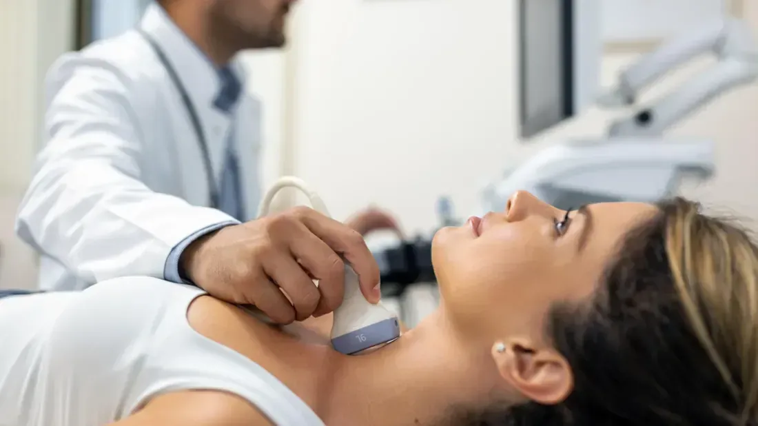 A male doctor in a white coat checking the blood pressure of a seated male patient in a plaid shirt.