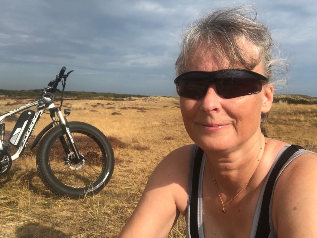 A portrait of Sally, a smiling woman in her 40s with auburn hair, wearing a linen apron, seated at a wooden workbench surrounded by bookbinding tools and pastel-coloured papers. The setting is bright and welcoming.