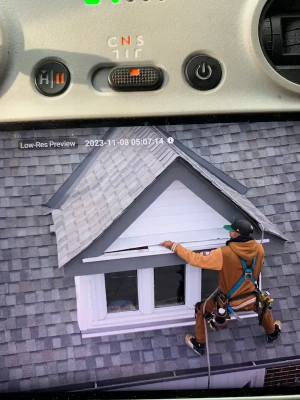 A-OK Roofing professional in a safety harness performing a detailed drone roof inspection on a Toronto residence. This high-angle view from their Precision Drone Inspections screen shows a harnessed technician assessing a dormer window and asphalt shingles. Critical for East York roof repairs and GTA drone roof inspections.