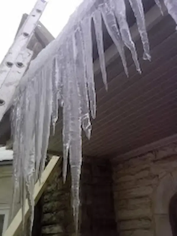 Severe winter ice damming with massive icicles hanging from a residential roof eave and soffit in Toronto. A roofing ladder rests against the house, demonstrating A-OK Roofing's rapid response for emergency leak repair. Critical visual for resolving winter storm damage and stopping active roof leaks for homeowners in East York and the Greater Toronto Area.