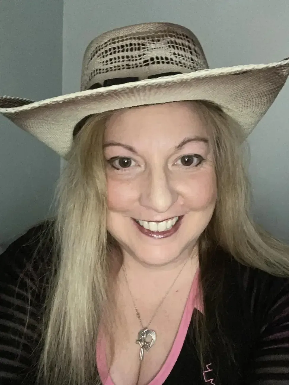A woman indoors is smiling and posing while wearing a cowboy hat.