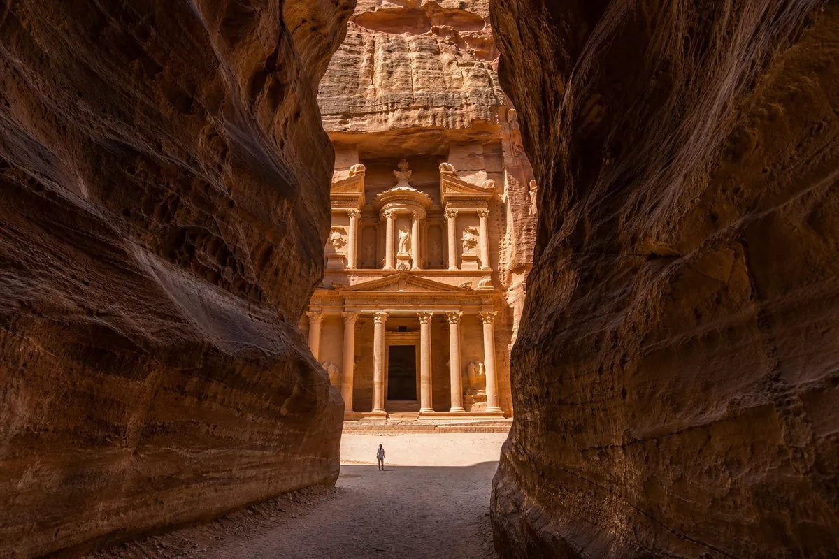 Narrow red sandstone canyon walls opening onto sunlit carved rock facade in the distance