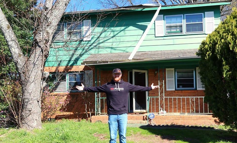 a man in a hoodie sweater standing in the grass with his arms raised in front of a green house with a porch