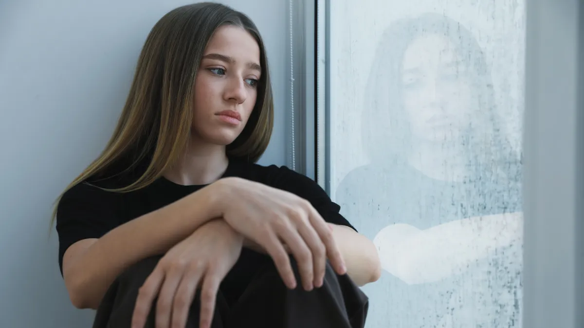 Pensive adopted teenage girl sitting by a window experiencing emotional struggles