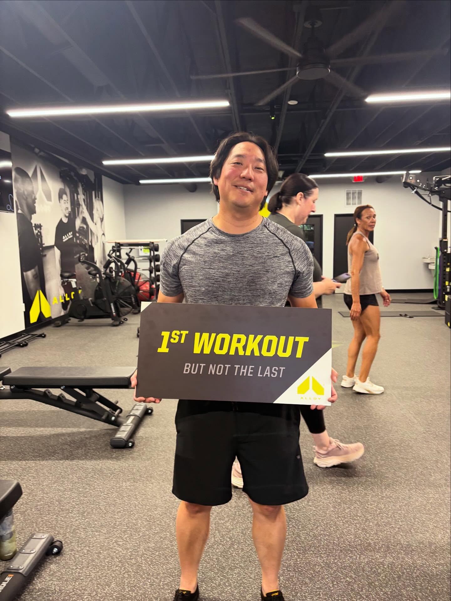 New client holds a sign celebrating their first workout at Alloy Personal Training, symbolizing their fitness journey in small group personal training.