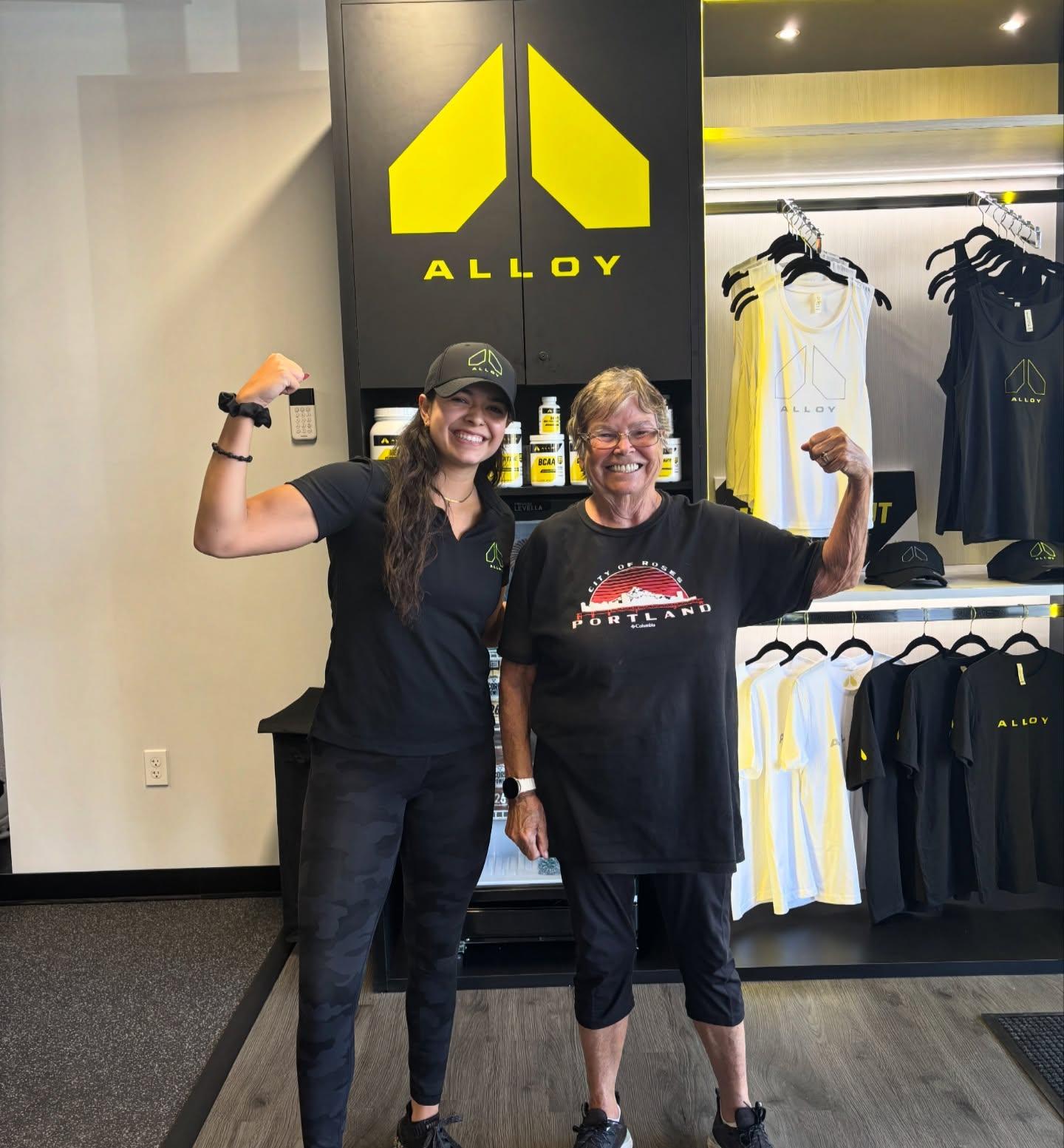 Personal trainer and client celebrating strength gains at Alloy Personal Training in front of branded gym apparel, showing progress in a strength training and functional fitness journey.