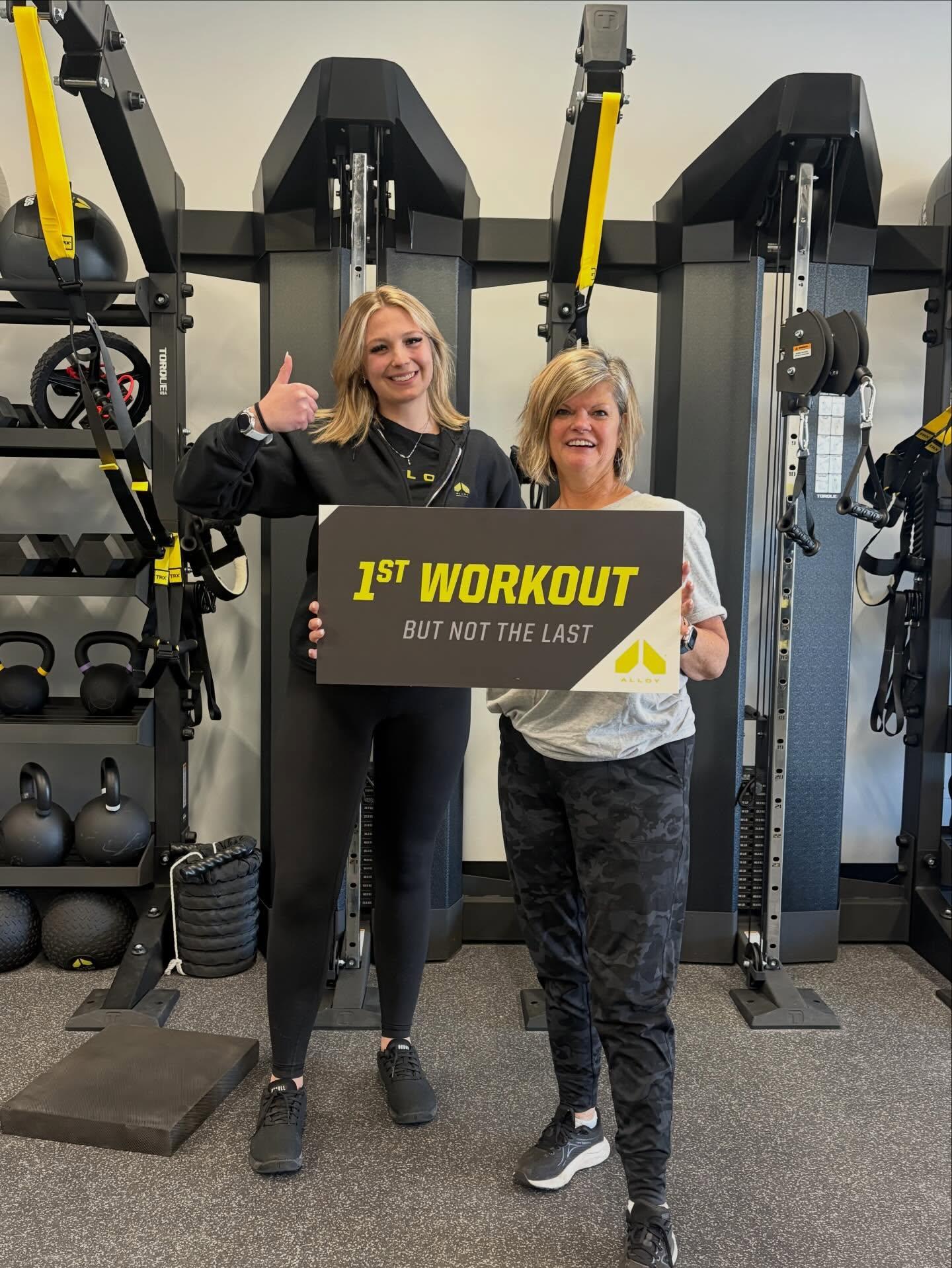 Client and personal trainer at Alloy Personal Training Geneva holding “1st Workout” sign in a small group training studio with strength training equipment, representing functional strength training and personal training in Geneva IL