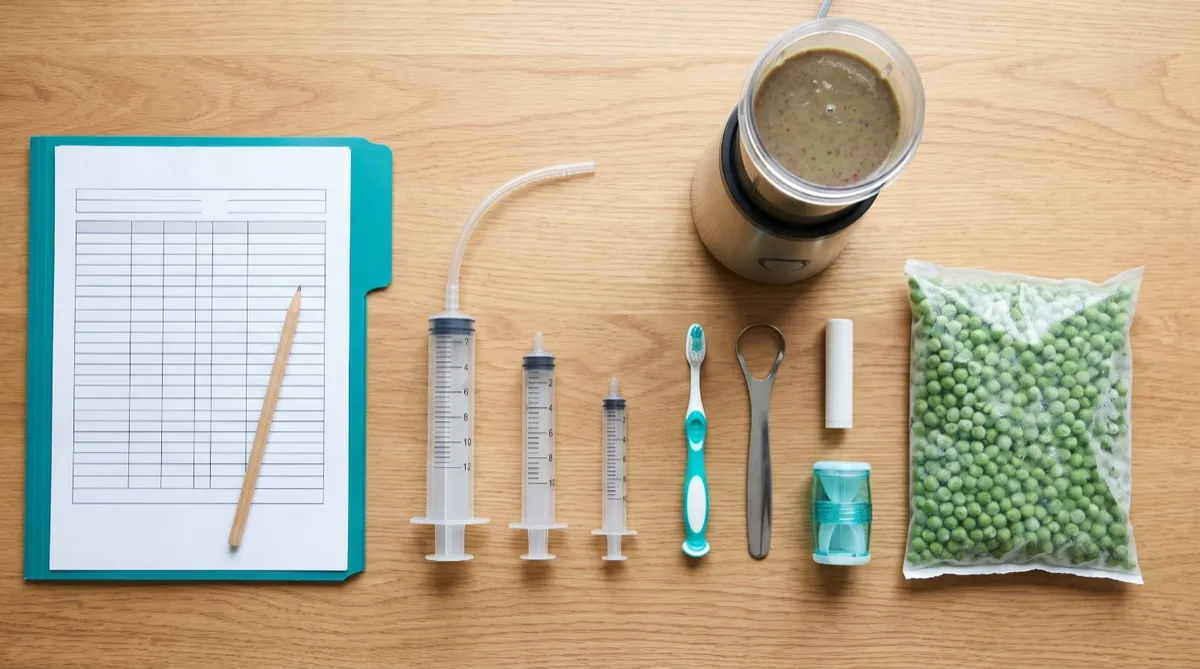A jaw surgery recovery kit on a wood counter: a teal folder with a chart, feeding syringes, a blender cup, a child toothbrush, a tongue scraper, lip balm and frozen peas.