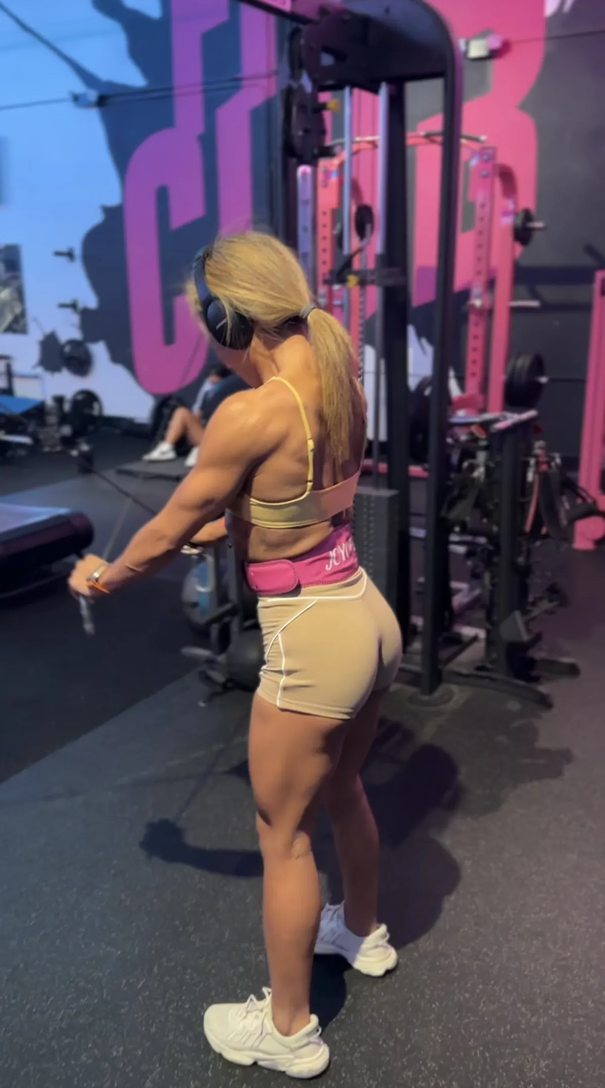 Picture of joy do a cross cable rear deal workout.  She is wearing a nude color sports bra and shorts and pink weightlifting belt.  