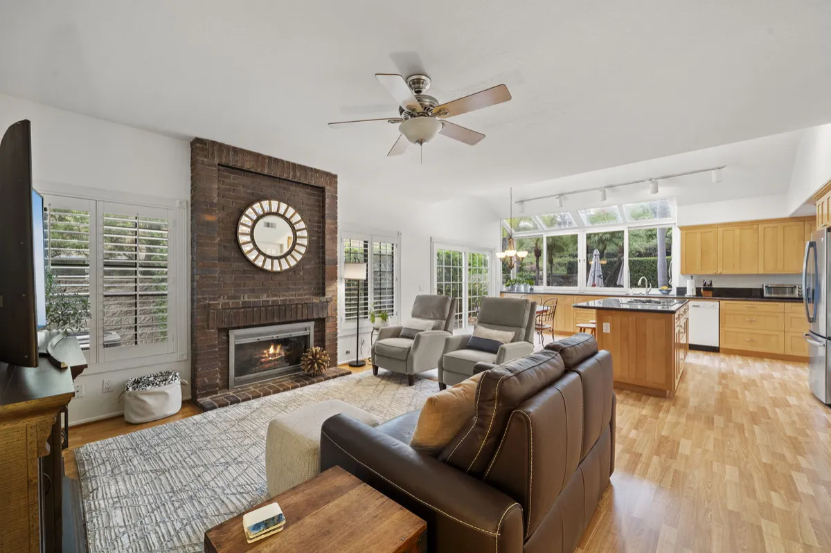 Family room with a brick fireplace, ceiling fan, leather sofa, and windows beside the adjoining kitchen.