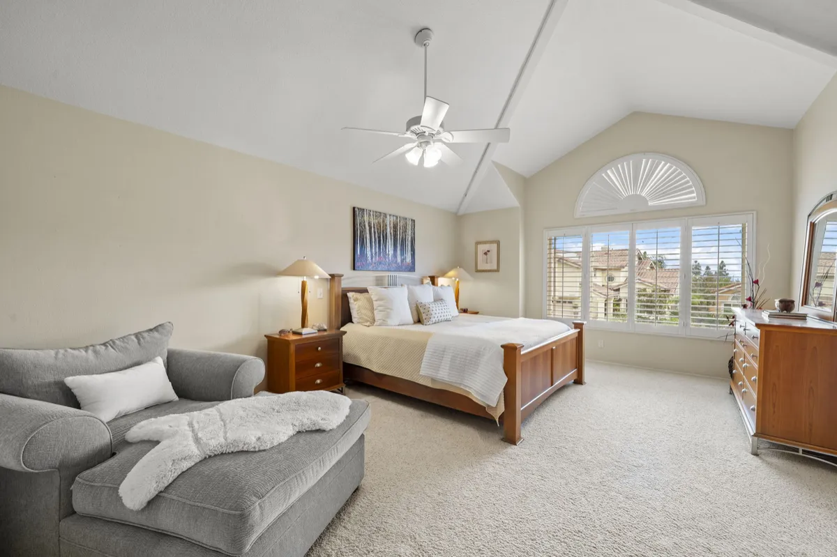 Primary bedroom with a wood bed frame, sitting area, ceiling fan, and plantation shutters beneath an arched window.
