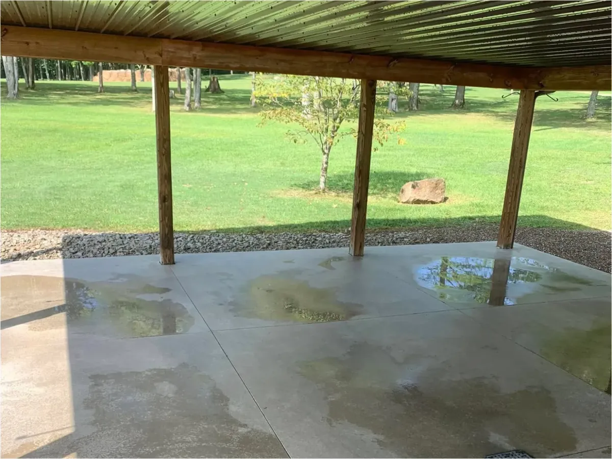 A concrete patio in an open space, with a patio cover. There are fresh puddles of water, showing signs the concrete has just been cleaned and restored. 