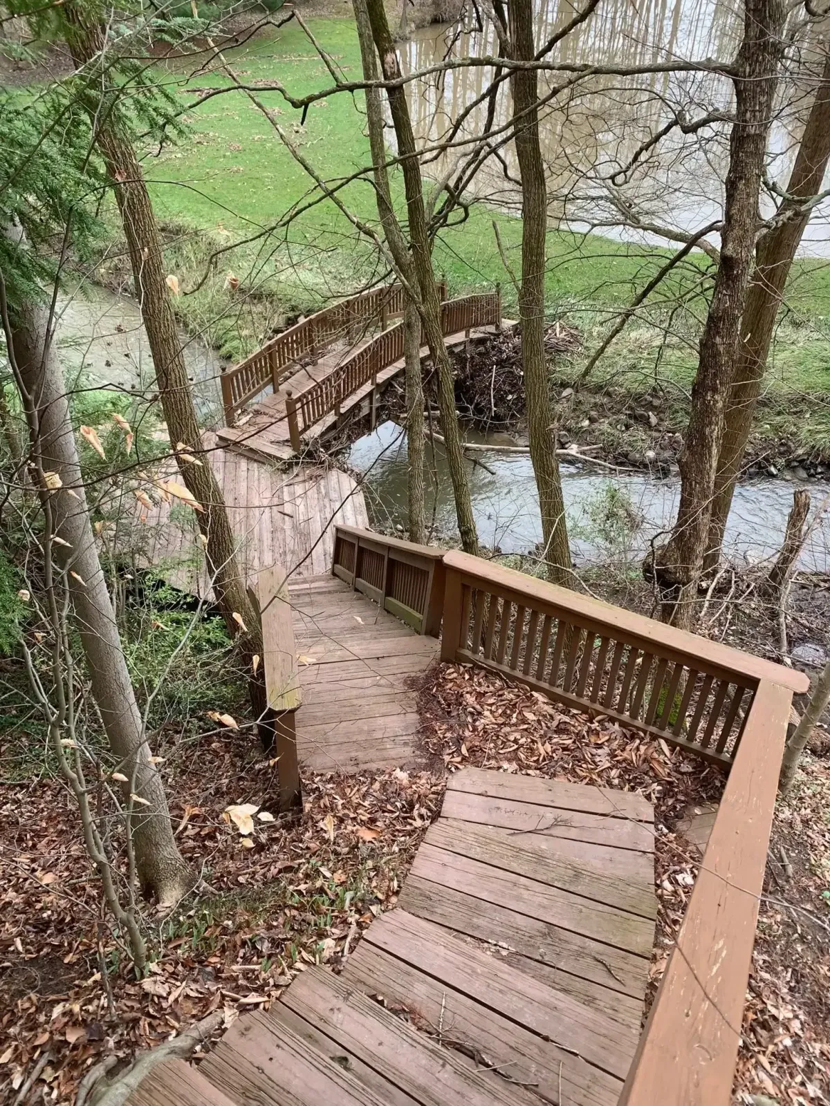 A long wooden pathway of a bridge leads over an open river. The pathway has lots of leaves, discoloration, and debris.