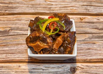 Stewed oxtail topped with julienned red, yellow, & green peppers served in a white rectangular bowl.
