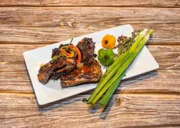 Jerk chicken drizzled with BBQ sauce sided with yellow & green habanero peppers and green onion served on a rectangular white plate.