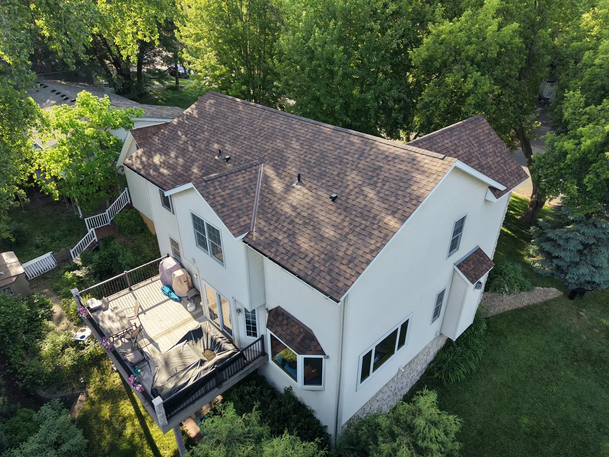 Drone view of a two-story white home with a brown architectural shingle roof, surrounded by mature trees