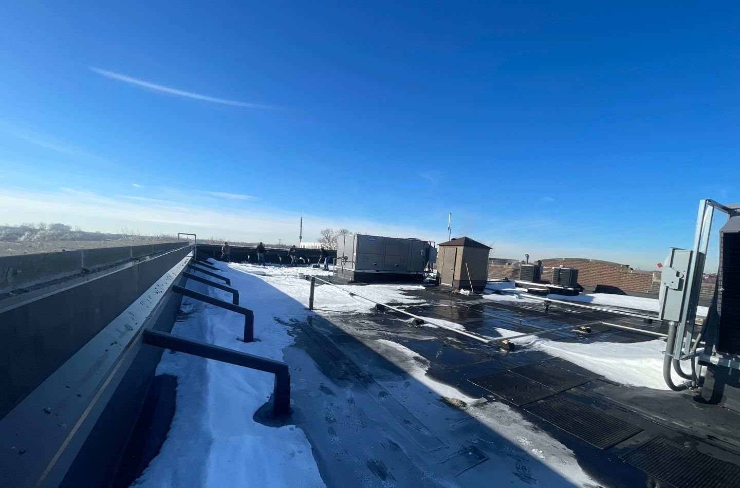 Flat commercial roof with snow packed along the parapet wall, partly cleared membrane, and rooftop units in the background