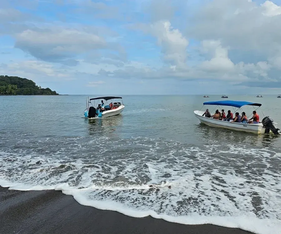 Wide tropical beach with dark sand, gentle waves, two passenger boats on calm water, and a lush green island beneath a partly cloudy sky.