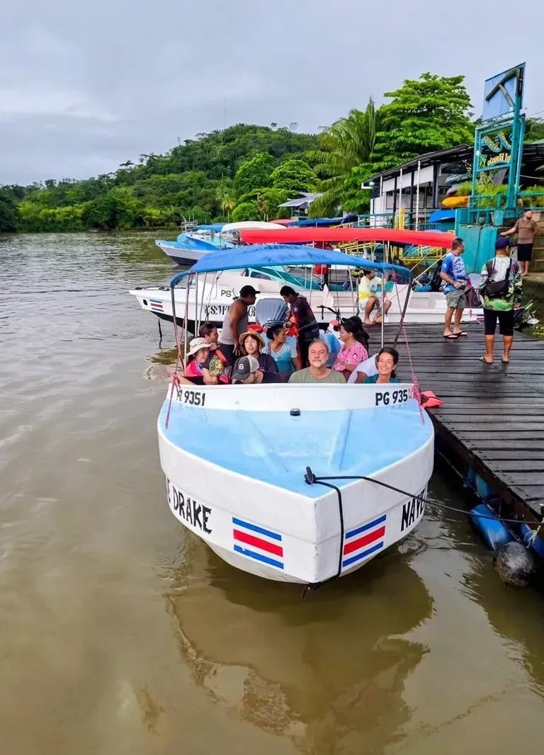 White and blue passenger boat with the Costa Rican flag, carrying people near a dock surrounded by lush green forest and other boats.
