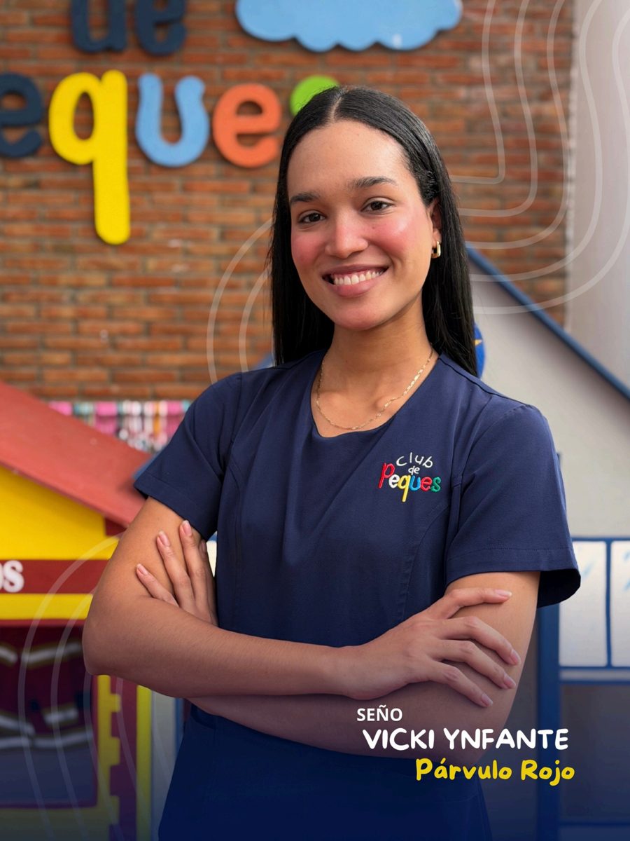 Waist-up portrait of Seño Vicki Ynfante with crossed arms, wearing a navy polo, in front of a colorful 'Peques' sign.