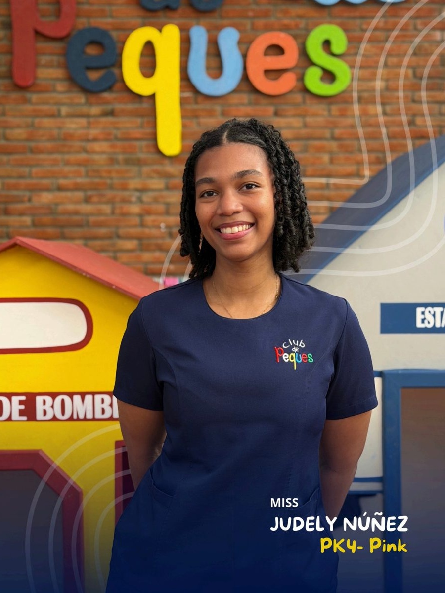 Waist-up portrait of Miss Judely Núñez smiling, wearing a navy polo, in front of a colorful 'Peques' sign.